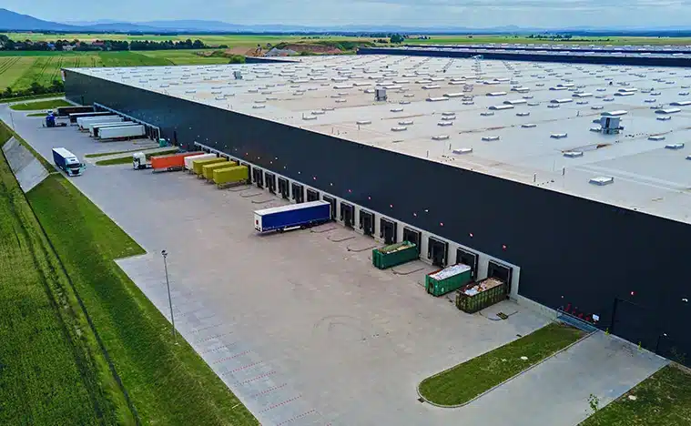Large modern warehouse with loading docks and parked trucks surrounded by green fields. Industrial logistics hub. Transport infrastructure for distribution, shipping and storage in supply chain system